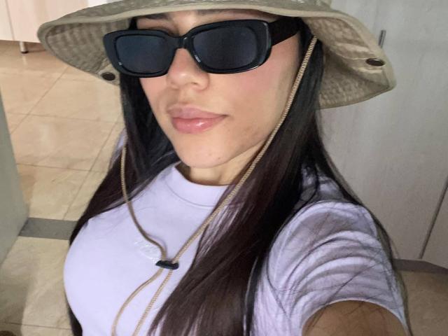 SophiaMarx with long dark hair, wearing a green bucket hat, black sunglasses, white t-shirt, and gold necklace, showing her slightly parted glossy lips and small to medium tits.