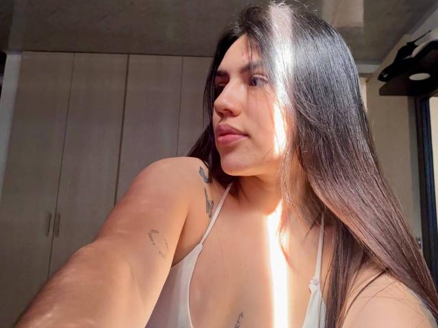 SophiaMarx with long dark hair, sunlit face, small tattoos on arm, white halter top, soft smile, light skin, taking selfie, visible boobs, natural light, calm expression.