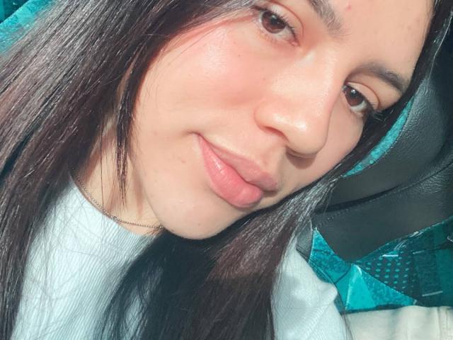 SophiaMarx's face, with smooth light skin, brown eyes, and dark straight hair, showing a subtle smile, wearing a white shirt, and sitting in a black car seat.