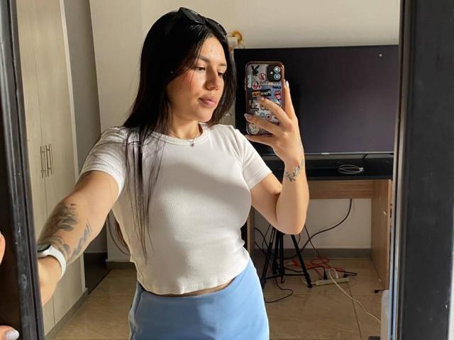 SophiaMarx with long black hair, white t-shirt, and blue pants, taking a mirror selfie showing her tits, small tattoo on her right arm, and holding a phone with stickers.