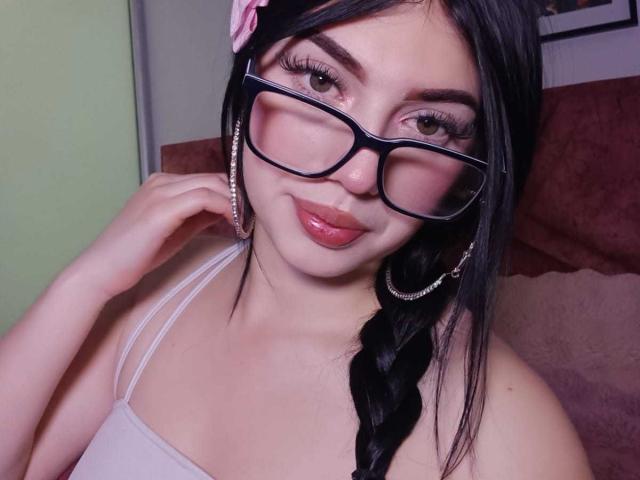 Isabella Rossi, con capelli neri intrecciati, occhiali, un fiore rosa, orecchini a cerchio, labbra rosa lucide, canotta bianca, che tocca la guancia, sorridente, pelle chiara, occhi marroni, sfondo rosa-rossastro..