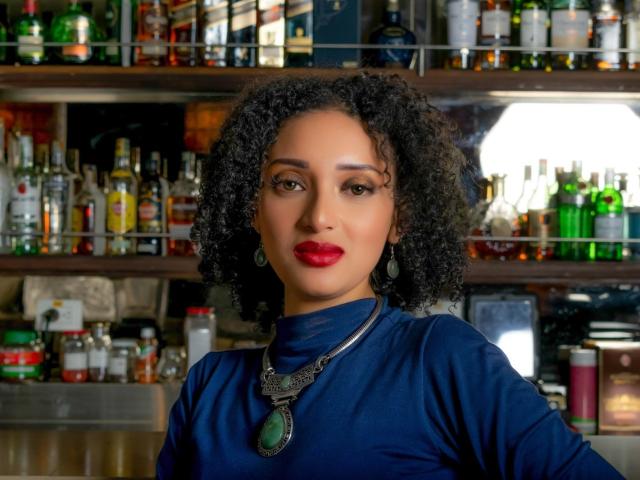 VioletaSwitch with curly black hair, red lipstick, blue dress, green necklace, and green earrings, standing in a bar, surrounded by liquor bottles.