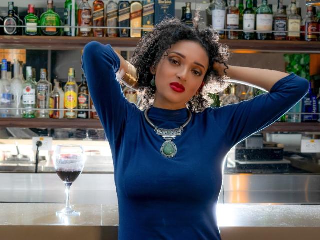VioletaSwitch with curly black hair, red lipstick, blue long-sleeve top, necklace, hands in hair, standing at bar, glass of red wine on counter, shelves of liquor in background.