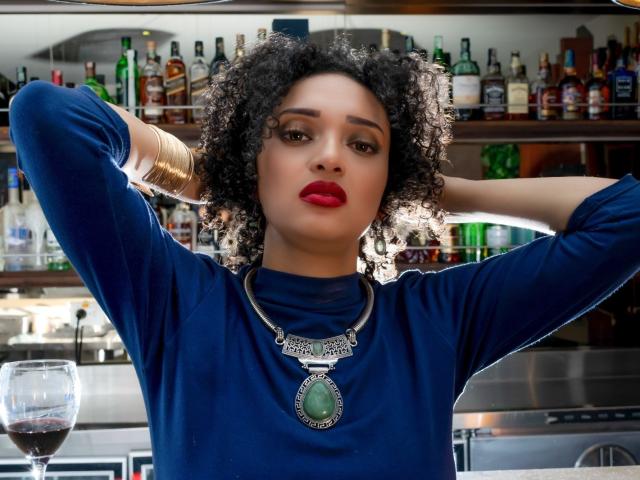 VioletaSwitch, con cabello negro rizado, lápiz labial rojo, vistiendo una blusa azul de manga larga, una pulsera dorada y un colgante verde, con los brazos levantados, las manos detrás de la cabeza, y una copa de vino delante..