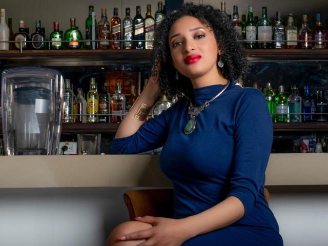 VioletaSwitch with curly black hair, red lipstick, and blue dress, sitting at bar, wearing gold bracelet and necklace, hands on knee, looking at camera.