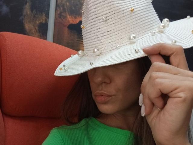 ViktoriiaLu wearing a white straw hat with pearl buttons, pouting with lips slightly pursed, green shirt, long brown hair, holding hat with right hand, red chair.
