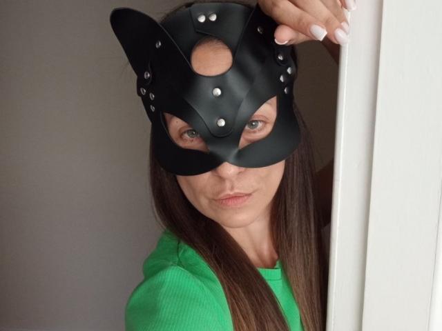 ViktoriiaLu with a black leather cat mask, green hoodie, and long brown hair, peering from a white door, showing her blue eyes and pink lips.