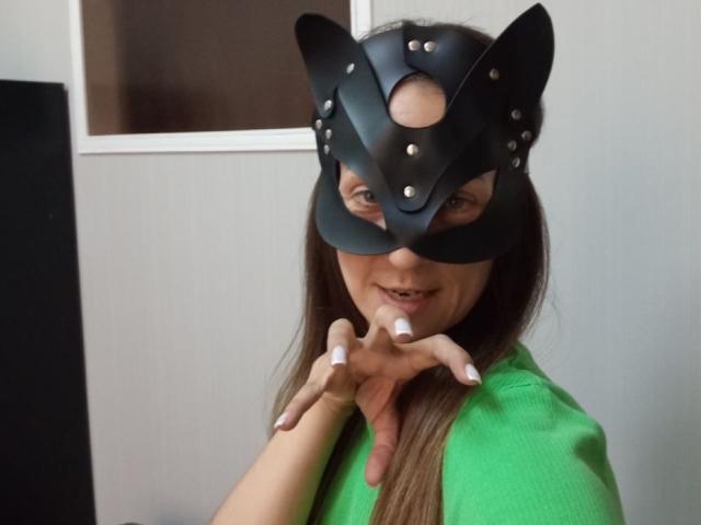ViktoriiaLu wears a black leather cat mask with metal studs, smiling with her hands near her mouth, green shirt, long brown hair, and white nails.