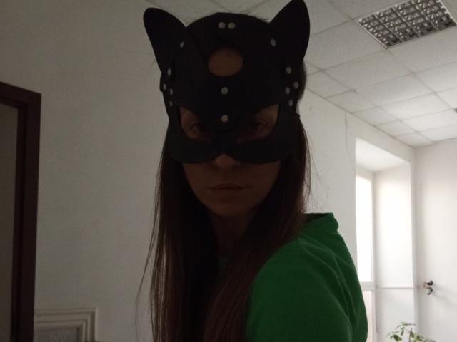 ViktoriiaLu with long brown hair, wearing a black cat mask with ears and holes, and a green hoodie, in a dimly lit room, looking down.