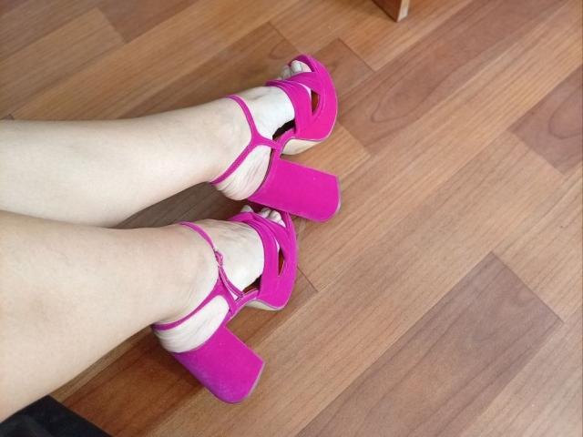 ViktoriiaLu's pale legs in bright pink high heels with chunky platforms, standing on a wooden floor. Her feet are slightly apart, showcasing the vibrant shoe color.