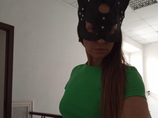 ViktoriiaLu wearing a black mask with holes, green shirt, and long brown hair, showing her big boobs, in a dimly lit room.