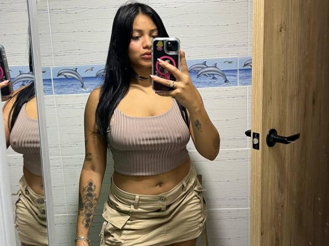 AlanaLuxe mit langen, schwarzen Haaren, einem braunen, gerippten Crop-Top, das ihre Brüste zeigt, einem beigen Rock, Tätowierungen am rechten Arm, hält ein Telefon in einem Spiegel-Selfie, eine Holztür auf der rechten Seite..