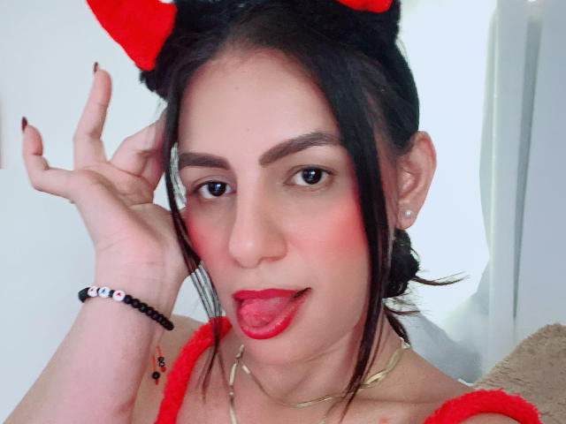 CamilleRiver has dark hair, red lips, and is sticking out her tongue. She wears red furry cat ears, a red top, and a black bead bracelet.