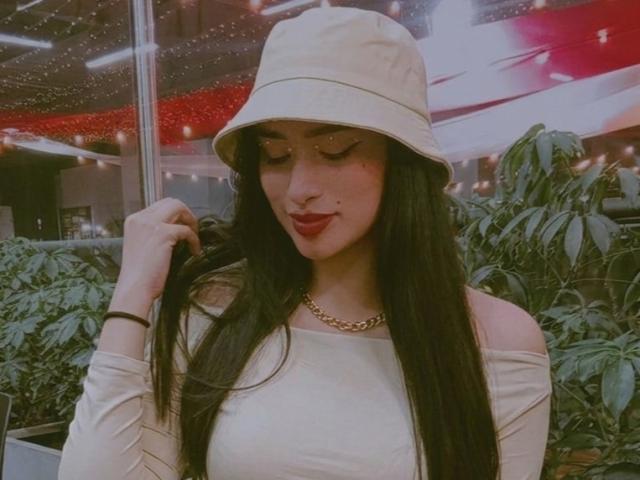 Adalina Vera, con cabello negro largo, usando un sombrero tipo cubo color beige y una blusa con un hombro descubierto, una cadena dorada, lápiz labial rojo, tocándose el cabello, plantas verdes y luces rojas en el fondo..