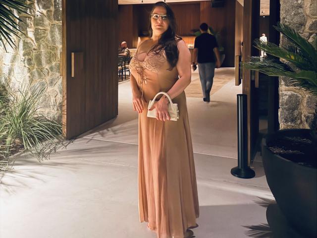 Ein Bad Angel mit großen Brüsten, der ein langes, hellbraunes, durchsichtiges Kleid trägt, eine weiße Clutch hält und in einem Türrahmen steht, in die Kamera schaut, dunkles Haar, Brille, eine Steinmauer im Hintergrund..