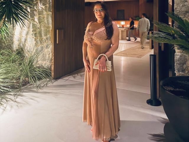 BadAngel com cabelos castanhos longos, óculos e seios grandes, vestindo uma blusa rosa de renda, um vestido marrom e saltos, segurando uma clutch branca, em um espaço moderno, iluminado pelo sol e cheio de plantas..
