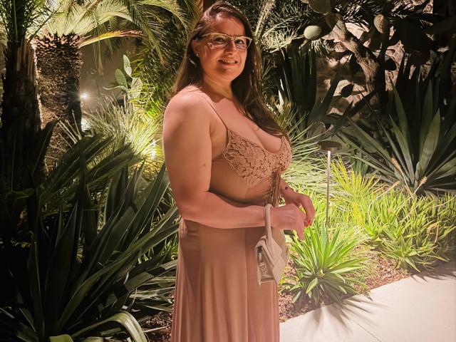 Ein BadAngel mit mittelgroßer Brust, braunen Haaren und Brille, trägt ein pinkes Spitzenkleid, hält eine weiße Tasche und steht in einem sonnendurchfluteten Garten und lächelt..