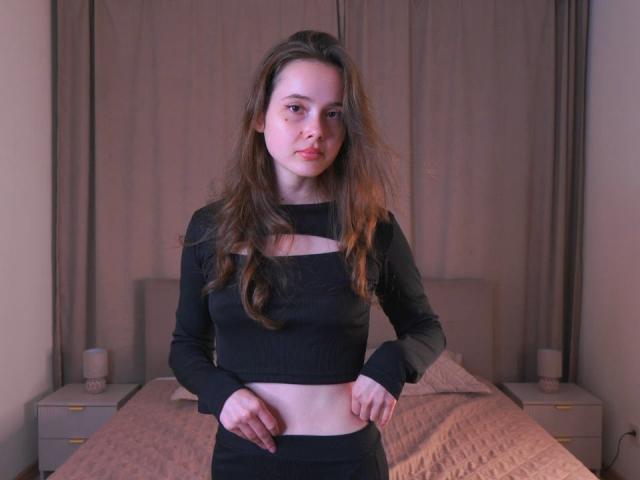 MeliaAmoroso with long brown hair, black crop top, and black skirt, standing in bedroom, hands on waist, showing her bare midriff and small ass.