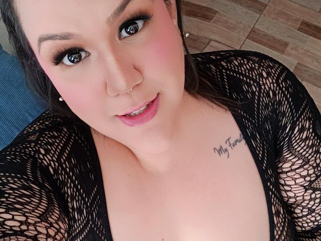 GiiannaMorrys with pink blush, black fishnet top, nose ring, and tattoo "My Girl" on her chest, looking up with dark eyes and pink lips. Boobs visible under fishnet.