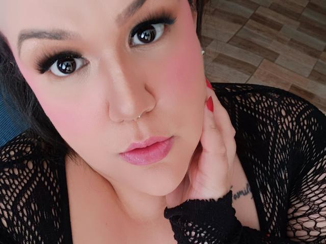 GiiannaMorrys with pink blush on her cheeks, black fishnet top, red nails, nose ring, and slightly parted pink lips, looking up with brown eyes, hand near her face.
