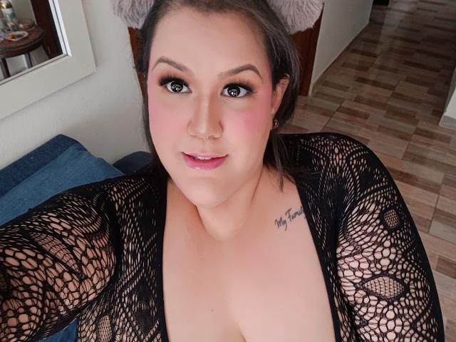 GiiannaMorrys with pink blush, black mesh top, small nose ring, tattoo on right chest, looking at camera, medium tits, brown hair, furry cat ears, wooden floor, blue pillow.