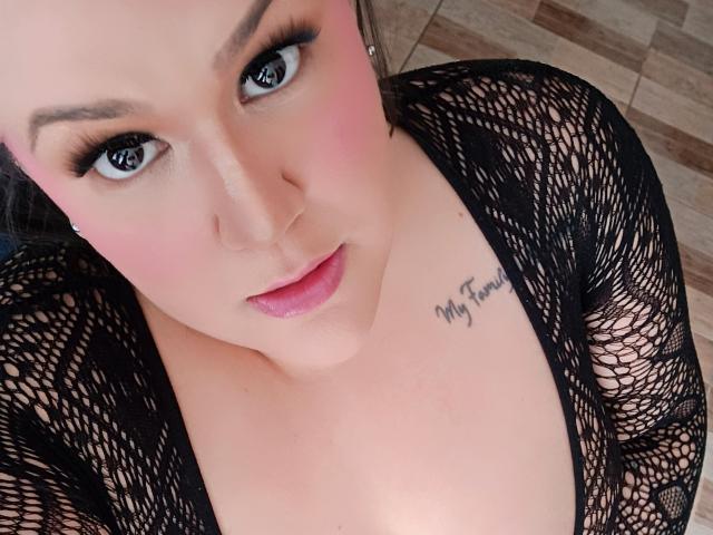 GiiannaMorrys with pink blush, black mesh top, blue eyes, pink lips, tattoo on chest, looking up, fair skin, visible cleavage, black hair, and subtle makeup.