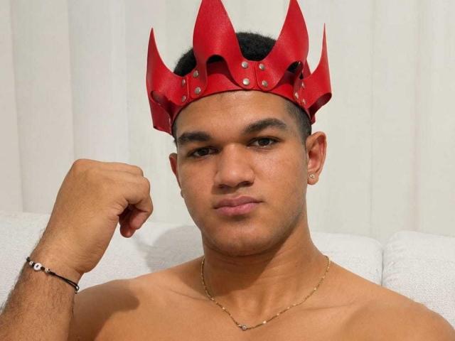 MaikolTwinkX with a red, spiked crown, shirtless, fist raised, black bracelet, gold chain, and earring, has a serious expression, dark skin, short hair, and small nose.