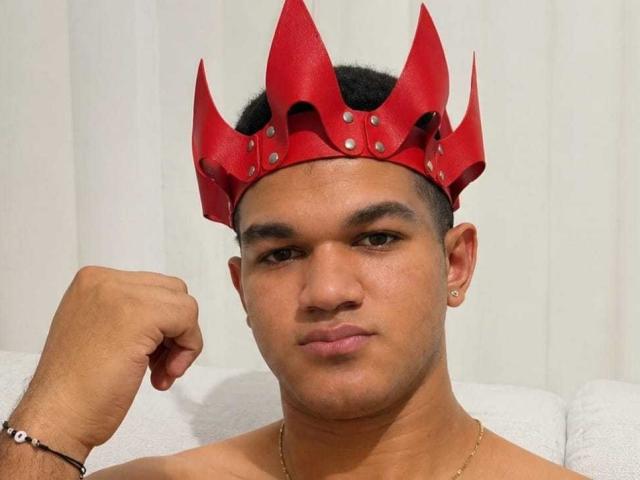 MaikolTwinkX with a red, spiked leather crown, fist raised, shirtless, tan skin, short black hair, small earrings, bracelet, and a neutral expression.