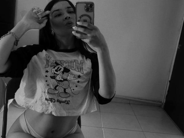 EmmaLombardi with dark hair, wearing a ragged Mickey Mouse shirt and panties, taking a mirror selfie, showing her flat stomach and slightly visible pussy, in black-and-white.