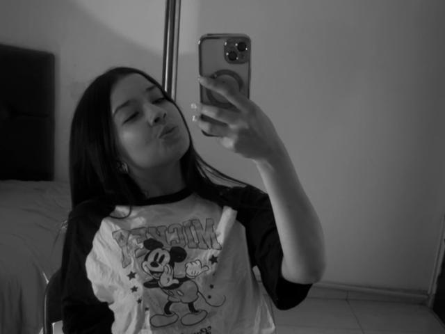 EmmaLombardi taking a selfie in a mirror, pouting with eyes closed, wearing a ragged Mickey Mouse shirt, holding an iPhone with a ring case, in a black-and-white photo.