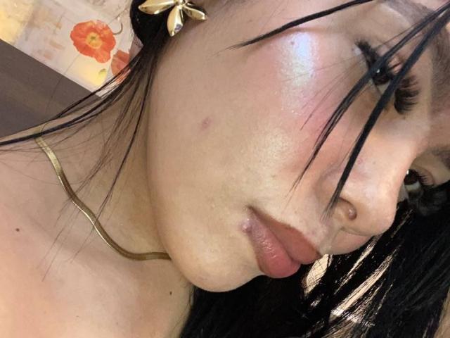 AdrianaPaul's face, showing glossy skin, slight acne, black hair, gold necklace, and a small lip piercing, with a flower hair clip, in a slightly tilted selfie.