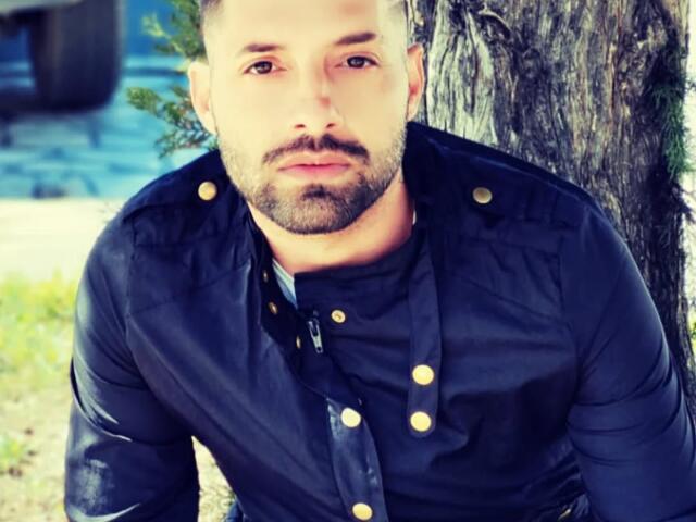 TommyDum with a beard, short brown hair, and serious expression, wearing a black shirt with gold buttons, leaning against a tree, slightly squinting eyes, and looking forward.