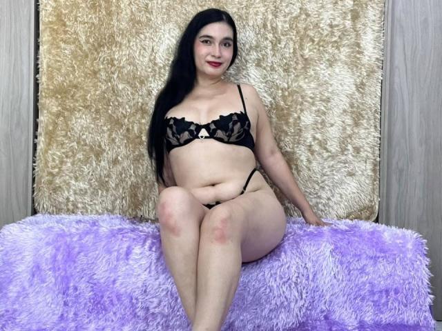 MaraLoop with black lace bra and thong, sitting on purple fuzzy blanket, long black hair, red lipstick, fair skin, slight belly, knees together, smiling, flabby ass.