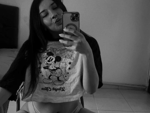 Black-and-white selfie of EmmaLombardi with long dark hair, pouting lips, wearing a Mickey Mouse shirt, and white panties, holding a phone in a tiled room.