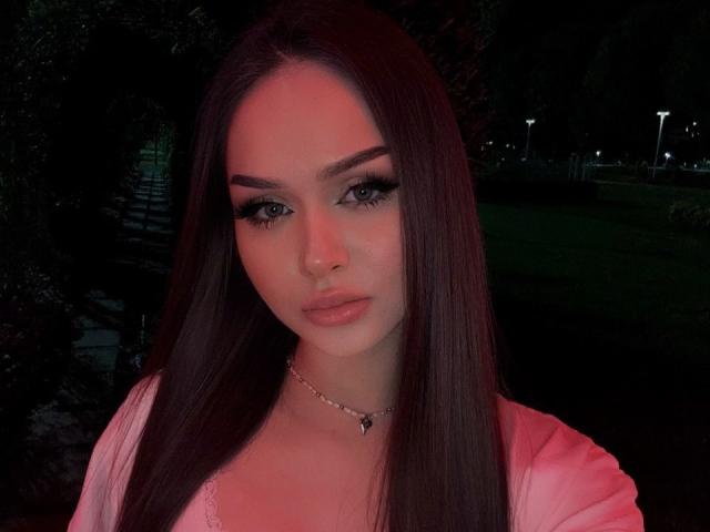 TamraBroxterman with long brown hair, pink lipstick, thick eyelashes, nose ring, and pink top, looking serious in a dimly lit outdoor setting. Her tits are small, visible under top.