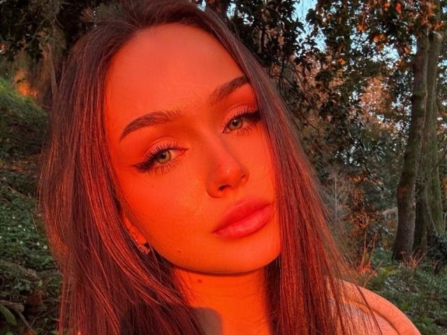 TamraBroxterman has orange-tinted skin, green eyes, long brown hair, and full pink lips. She's looking at the camera with a slight pout, wearing a nose ring.