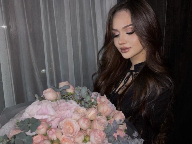 TamraBroxterman with long brown hair, wearing black, holding pink rose bouquet, looking down with a soft smile, standing by dark wooden wall, showing off her tits.