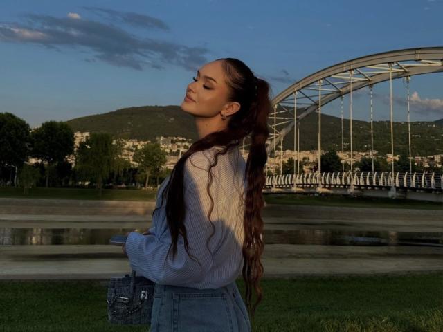 TamraBroxterman with long wavy brown hair, eyes closed, wearing a blue shirt and high-waisted jeans, standing by a river, bridge in the background, sunlight on her face.