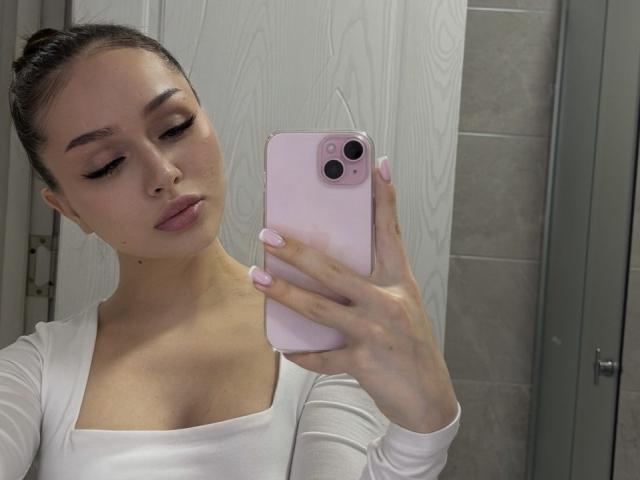 TamraBroxterman with pink nails, white top, and updo, holding pink phone, taking selfie in bathroom, showing off her boobs and a cute nose piercing.