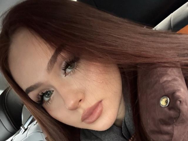 TamraBroxterman has red hair, green eyes, and full pink lips. Her eyebrows are thick, and she's wearing a grey shirt with a brown jacket. Her eyelashes are long.