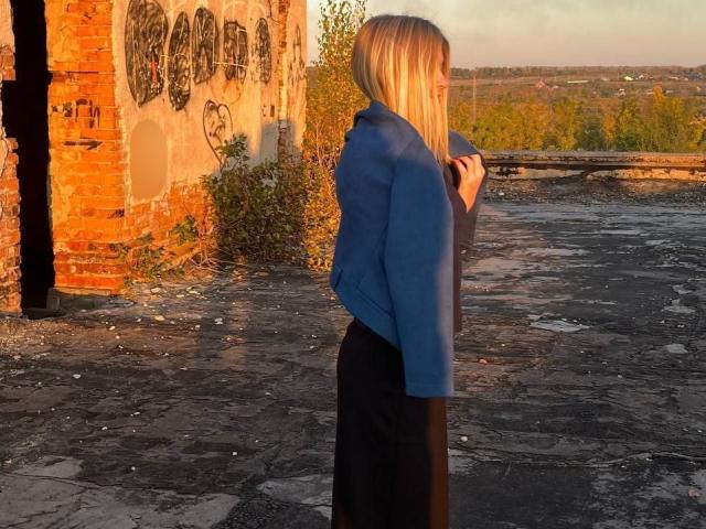 Norah Habrock è in piedi in un edificio in rovina, illuminato dal sole, indossa una giacca blu sopra una gonna nera, ha i capelli biondi, tiene la giacca in mano, mostrando il sedere, e ha un'espressione triste..