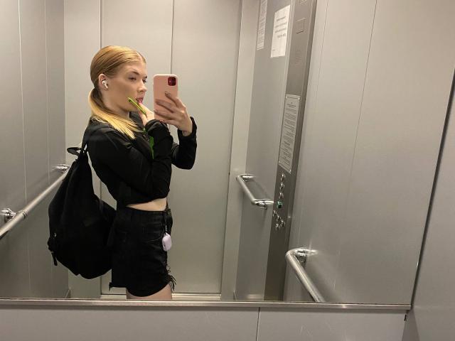 NorahHabrock, blonde, dans un ascenseur, prend un selfie devant un miroir, portant une veste noire, un haut vert, un short noir, un sac à dos, des écouteurs, un téléphone rose, vue de profil, petite poitrine, mince, mignonne, se reflétant sur les murs métalliques..