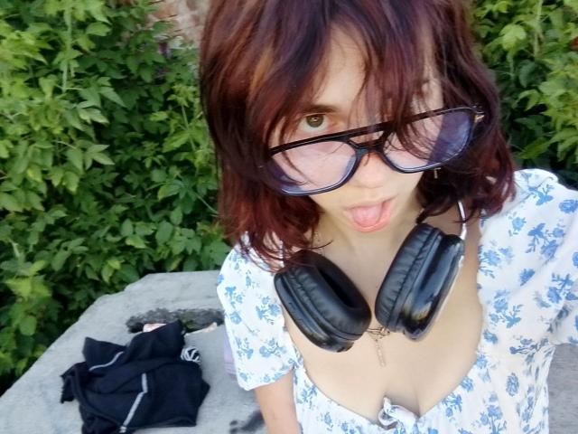 Jenice Arrellano, con cabello castaño rojizo, gafas y auriculares negros, haciendo pucheros, vestida con un vestido blanco con estampado floral, mostrando su escote, un bolso negro sobre una superficie gris, fondo verde..
