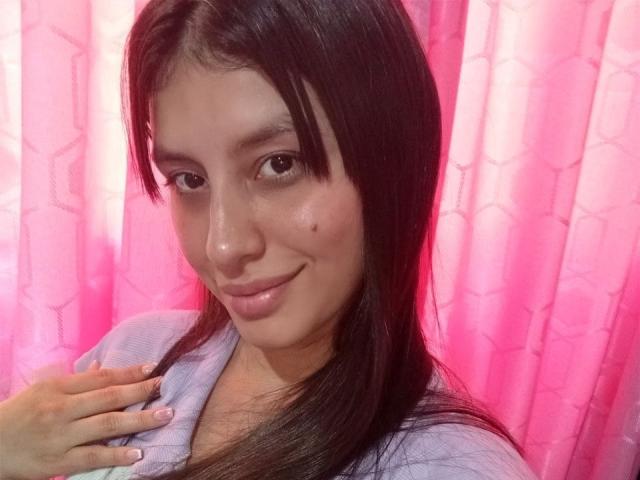 MadissonFontaine with long black hair, light skin, subtle smile, small mole on cheek, wearing light purple top, hand on chest, pink curtains, soft pink lighting, gentle expression.
