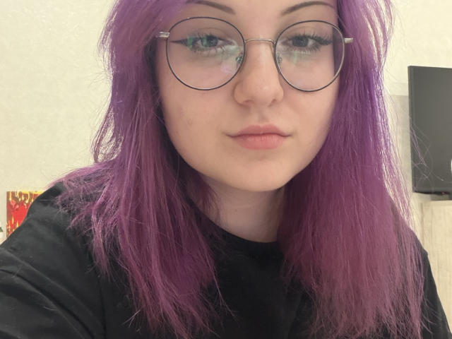 StevieRerucha, con cabello morado, gafas redondas y una camisa negra, muestra una expresión neutra. Sus senos no son visibles, solo su rostro. Está mirando directamente a la cámara..