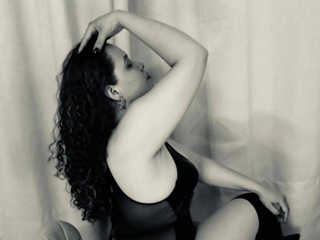 VittoriaI with curly dark hair, wearing a black sheer top, sits with arm raised, hand in hair, showing her tits, against a neutral background, in a black-and-white photo.
