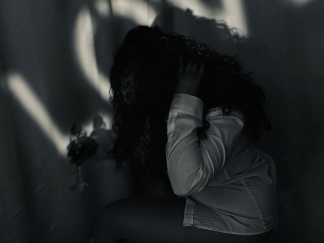 VittoriaI has dark curly hair, wearing a white shirt, sitting with hands in hair, shadowy light patterns on her face, her ass slightly visible, in a dark, moody photo.