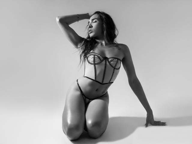 GabySantos is kneeling, wearing a strapless, bejeweled corset and matching thong. She's touching her head, eyes closed, with a hand on the floor. Her ass is visible.