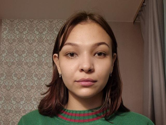 MairaMoctezuma has brown eyes, straight brown hair, and wears a green sweater with red stripes. She has small earrings and a neutral expression. Her skin is light brown.