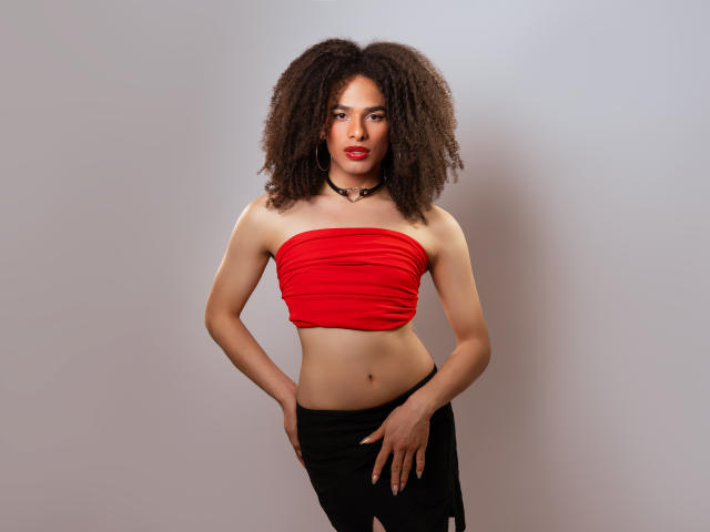 KataleyaFowler with curly brown hair, red tube top, black choker, and black skirt, standing with hands on hips, red lipstick, slim build, and a confident expression.
